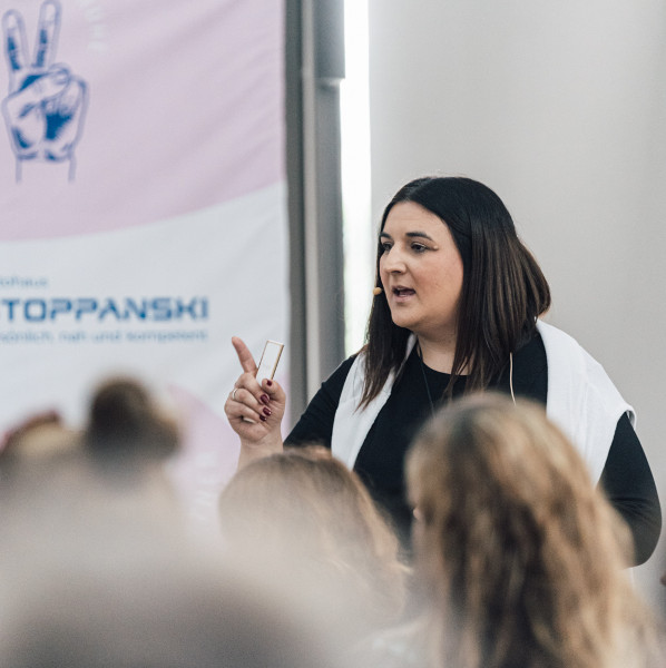 Eine Frau steht vor einem Publikum und gestikuliert mit erhobenem Zeigefinger, während im Hintergrund ein Banner mit dem Logo "TOPPANSKI" zu sehen ist.